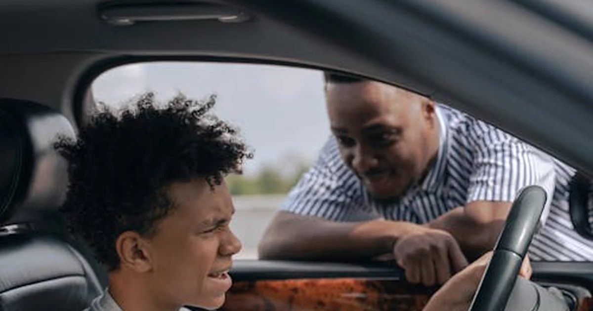 Adult instructor coaching a teenage student driver from the passenger seat during a sunny first lesson